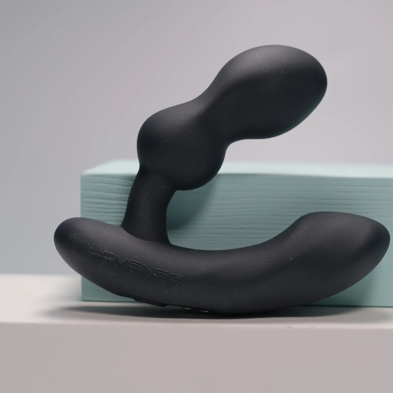 Lovense Edge 2 black app-controlled prostate massager displayed on a white surface in front of a pale mint block against a soft grey background.
