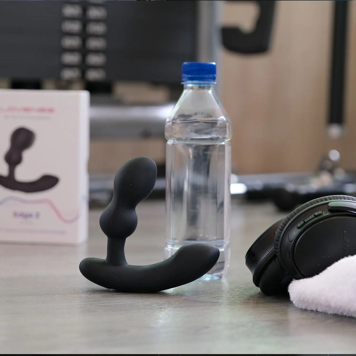 Lovense Edge 2 black prostate massager standing upright on a tabletop beside a water bottle, with headphones and product packaging in the background.