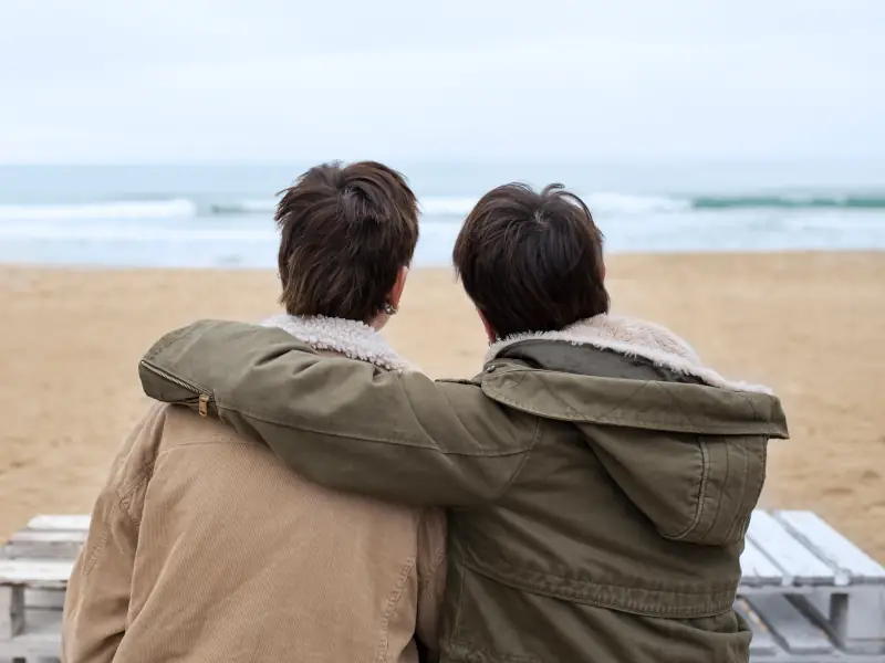 Two people in jackets sitting together on a beach and looking out at the sea